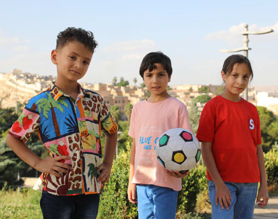 Three children stand outdoors on a sunny day. The child on the left wears a colorful printed shirt, the middle holds a soccer ball, and the right wears a red shirt, all smiling against a blurred cityscape backdrop.