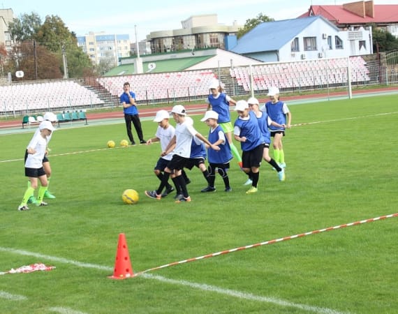 Kids in white and blue jerseys play soccer on a grassy field, surrounded by orange cones. A coach observes from the sidelines.