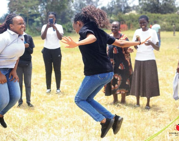 A group of people are outside on a grassy field. Two women are jumping rope while others stand around, watching, smiling, and taking photos. The mood is lively and joyful.