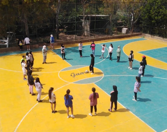 A group of children wearing casual sports clothes stand in a circle on a colorful outdoor court, with a soccer ball in the center.