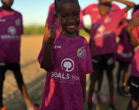 A smiling child in a pink soccer jersey makes a peace sign on a sunny day, with other teammates in matching jerseys in the background.