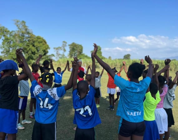 A group of children standing in a circle on a grassy field under a clear blue sky, holding hands with arms raised.