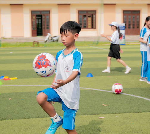 A young boy in a blue and white sports uniform skillfully balances a soccer ball on his knee on a grassy field, with classmates playing in the background.