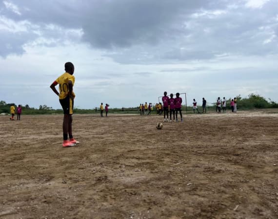 A boy in a yellow jersey stands near a soccer ball on a dirt field, preparing to kick while a group of kids in purple shirts form a defensive wall in front of a goal under a cloudy sky.