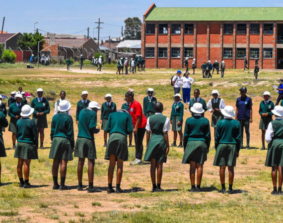 A group of students in green uniforms stand in a circle on a grassy field, with others scattered around and a large brick school building in the background. The weather is sunny and bright.