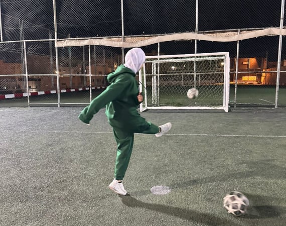 A person wearing a green tracksuit and white headscarf kicks a soccer ball on an outdoor field at night, with a goal and a chain-link fence in the background.