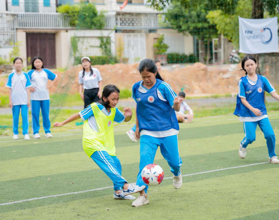 Two girls, one in a yellow vest and another in a blue vest, are playing soccer on a grassy field, focused on the ball. There are other players and observers in the background.