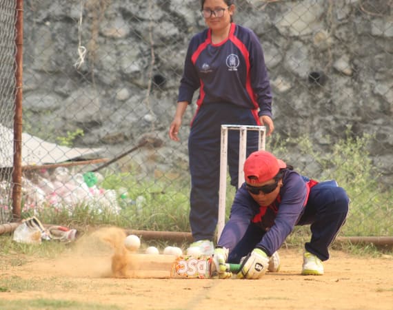 A cricket player in a red cap and sunglasses dives to catch the ball, while another player in matching gear watches. Dust rises from the ground, showing motion.