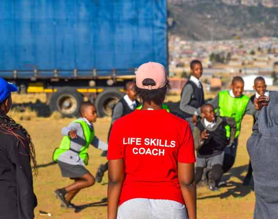 A woman wearing a red shirt labeled LIFE SKILLS COACH watches a group of schoolchildren in uniforms and green vests playing outdoors, with a blue truck and town in the background.