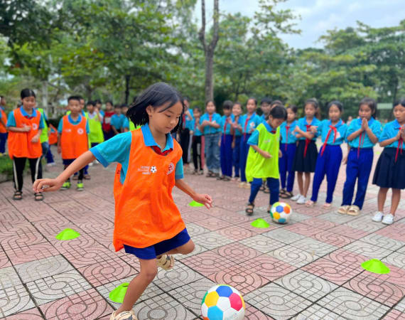 Two young girls one an orange vest and another in neon green vest dribble a colorful soccer balls around cones on a tiled playground. Their classmates watch, smiling and clapping, wearing blue uniforms.