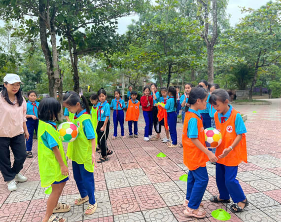 Children in colorful bibs play with balls in an outdoor park. They are surrounded by trees and guided by an adult.