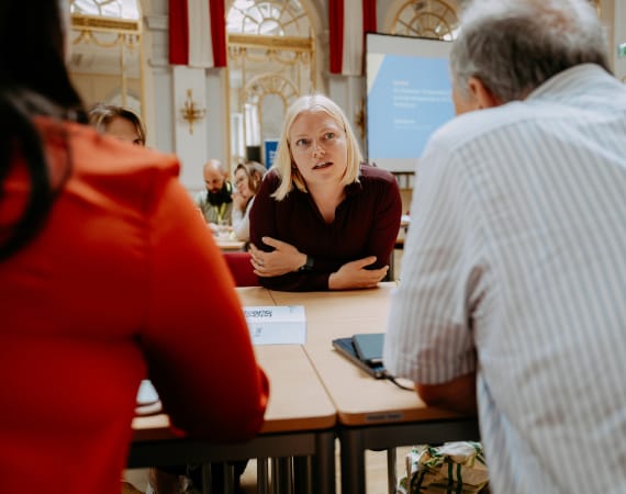 A woman in a maroon shirt leans forward, attentively listening to colleagues in a conference room. The scene conveys focus and engagement.