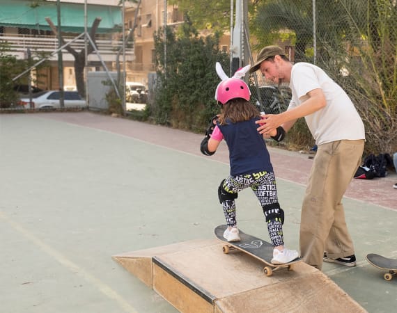 A child wearing a pink helmet with bunny ears skateboards down a small ramp. An adult is assisting her.