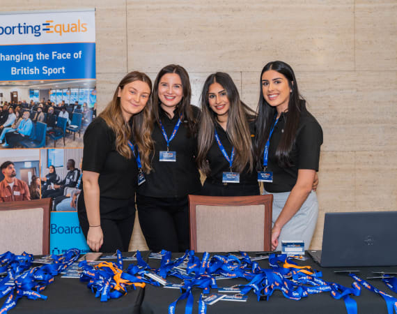 Four women wearing black shirts and name badges stand behind a registration table with lanyards, in front of a Sporting Equals banner at an event.