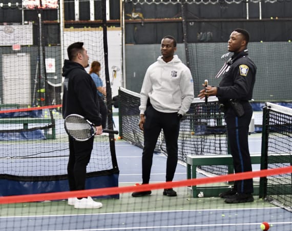 Three men stand on an indoor tennis court: one holding a racket, one in casual clothes, and one in a police uniform. They are talking near the net, with sports equipment and barriers in the background.