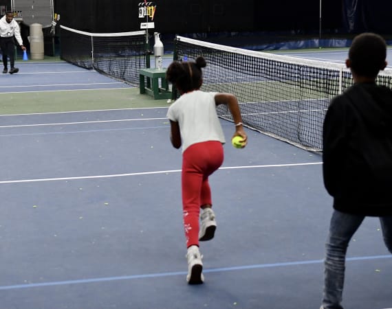 A young girl in red pants and a white shirt runs on an indoor tennis court holding a tennis ball, while another child walks nearby. A net and tennis equipment are visible in the background.
