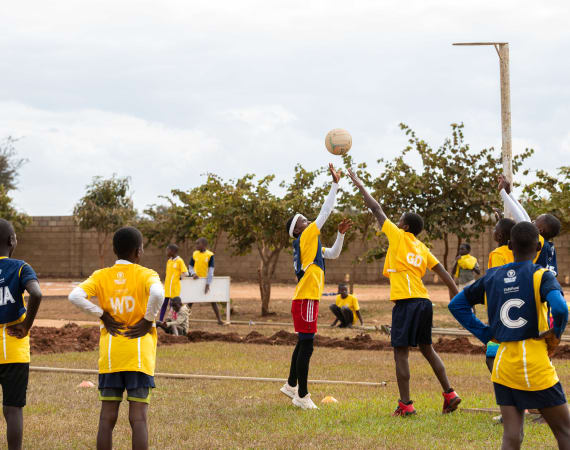 Children in yellow and navy uniforms play a lively netball game on a grassy field. Trees and a few onlookers are in the background under a cloudy sky.