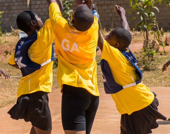 Children in yellow and blue sports uniforms jump and reach for a red ball on an outdoor court under a blue sky, with a brick wall and plants visible in the background.