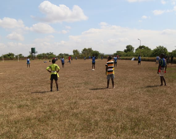 Children and teenagers play soccer on a dry grassy field under a partly cloudy sky, with some players wearing colorful jerseys and others watching from the sidelines. Trees and bushes border the field.