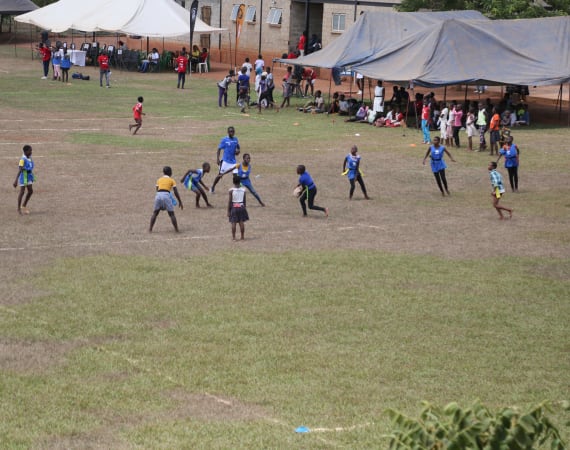 Children play a game on a grassy field, surrounded by people watching under large tents. Some are wearing blue shirts, while others wear different colors. In the background, more people gather near buildings.