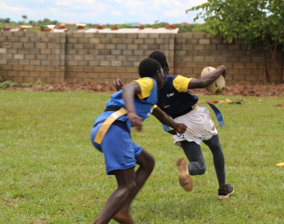 Two boys play tag rugby on grass. One, in a blue uniform, reaches to grab the flag belt of the other, who is wearing white shorts and running with a rugby ball. A brick wall and tree are in the background.