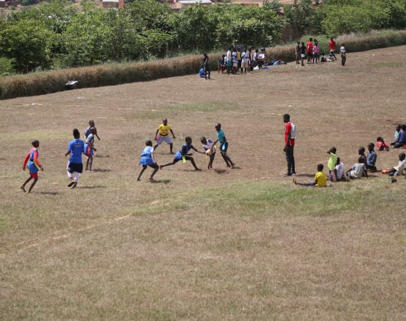 Children play a game of rugby on a grassy field, with some wearing colorful jerseys. A few adults and groups of people sit or stand nearby, watching the game. Trees and buildings are visible in the background.