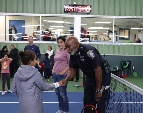 A police officer shakes hands with a young girl on an indoor tennis court, while other adults and children watch and interact in the background. A digital clock and glass windows are visible above the scene.