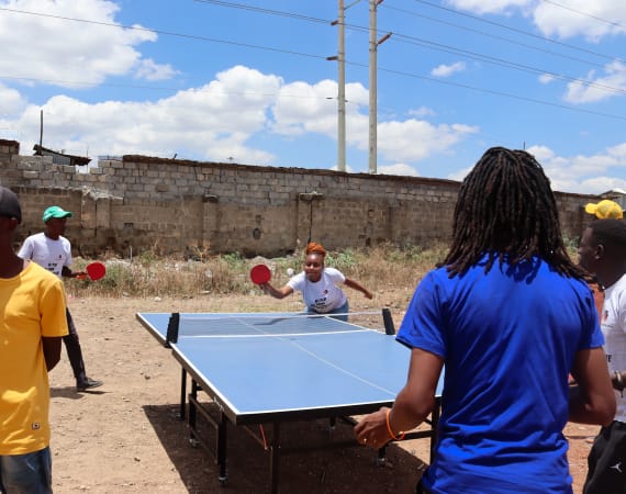 A group of people play table tennis outdoors on a sunny day, surrounded by a stone wall and power lines. One woman is mid-action, swinging her paddle, as others watch or wait their turn.