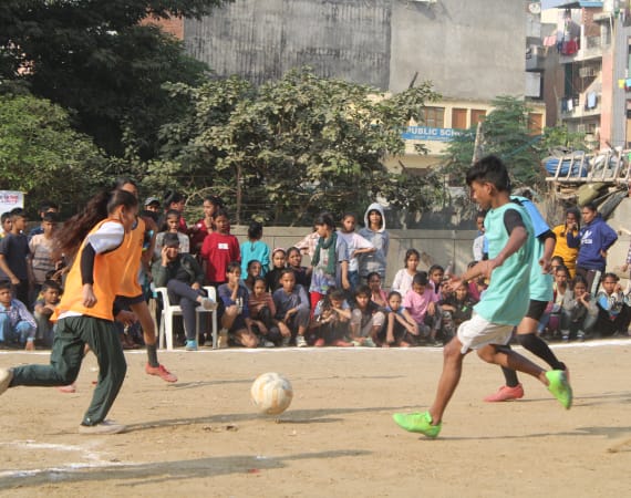 Children play a soccer match on a sandy field while a large crowd of spectators, including kids and adults, watch from the sidelines. Buildings and greenery are visible in the background.