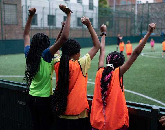 Three girls with braided hair, wearing bright sports vests, stand on the sidelines of a soccer field with their arms raised, cheering for players who are practicing in the background.