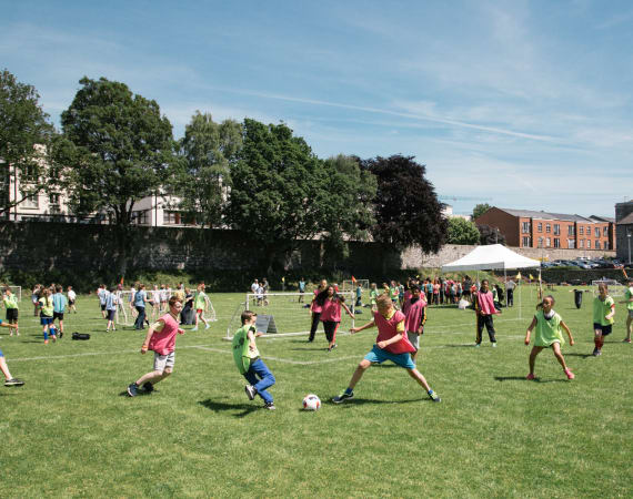 Children playing soccer on a grassy field during a sunny day, with other kids and adults in the background participating in different activities; trees, buildings, and a white tent are visible behind them.
