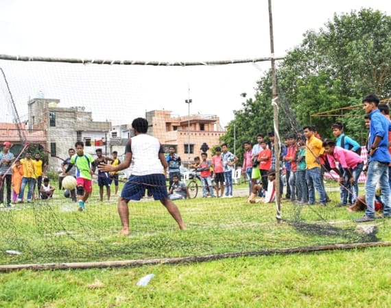 A group of people watch as a boy attempts to score a goal past a goalkeeper during an outdoor soccer game on a grassy field in a lively neighborhood.