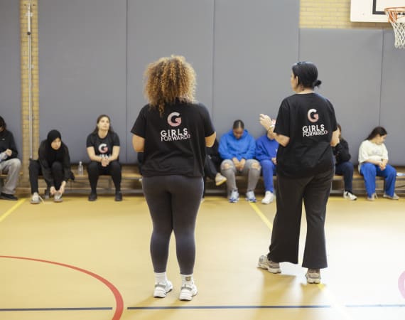 Two women stand in a gym, wearing "Girls Forward" shirts, facing a row of seated individuals.