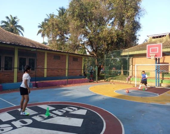 Two girls playing football basketball on an outdoor court surrounded by trees and a wire fence. The court has blue and red markings, and a brick building is visible in the background.