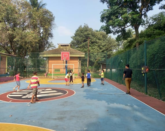 Children play basketball on an outdoor court surrounded by trees and a wire fence, while an adult walks nearby. The court has blue and red markings, and a brick building is visible in the background.
