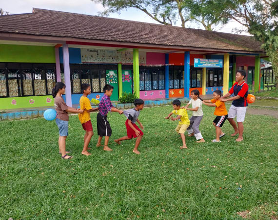 A group of children and one adult play a game in a grassy area in front of a colorful school building, holding onto each other in a line, smiling and laughing outdoors.