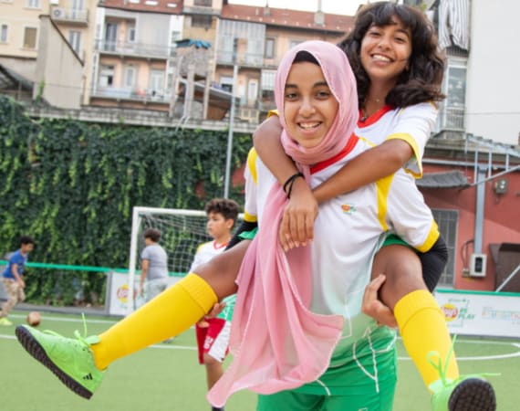 Two girls in soccer uniforms smile as one carries the other on her back on an outdoor field. Other children play in the background. Buildings and greenery surround the court.