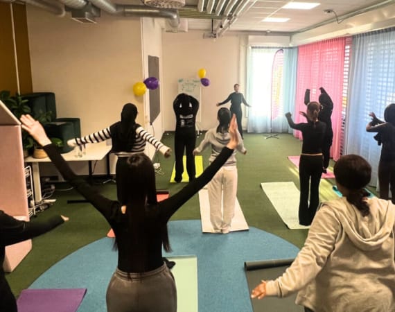 A group of people practice yoga or stretching on mats in a bright room with colorful curtains and balloons. Most have their arms raised. 