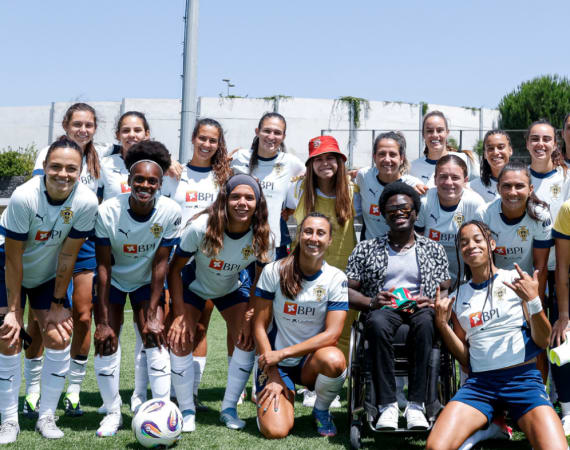 A women’s soccer team poses together outdoors on a grassy field, smiling at the camera. One person in a red hat and another using a wheelchair are in front.