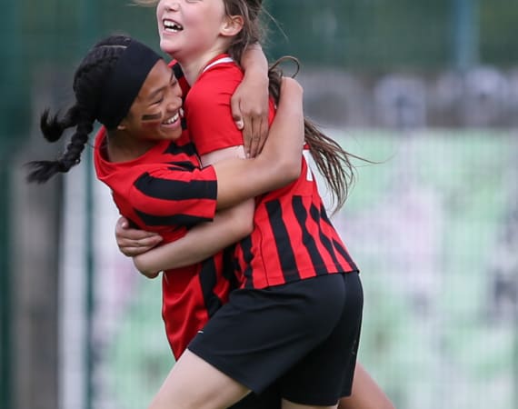 Two young girls in red and black soccer uniforms joyfully hug each other on a field, celebrating. One girl has face paint and braids.