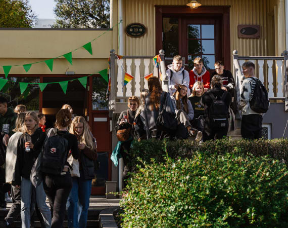 A group of students gathers outside a yellow building decorated with green and orange bunting. Some stand on the steps, others on the ground, talking and smiling in the sunlight.