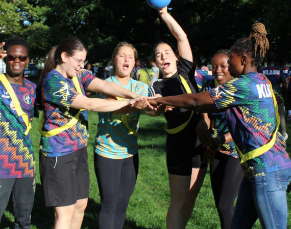 A group of people wearing colorful shirts stand in a circle outdoors, placing their hands together, with one person holding a blue ball aloft.