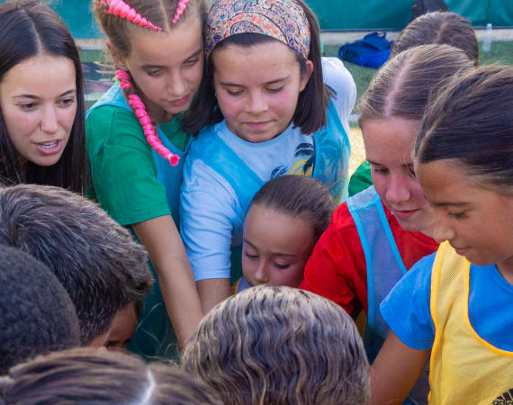 A group of young girls in sports jerseys huddle together with their hands stacked in the center, showing teamwork and unity at a soccer event.
