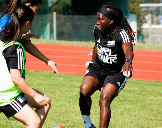 Three girls playing soccer on a grassy field. One girl is defending against two others as they go for the ball. All are wearing black athletic uniforms with GIRL POWER written on them.