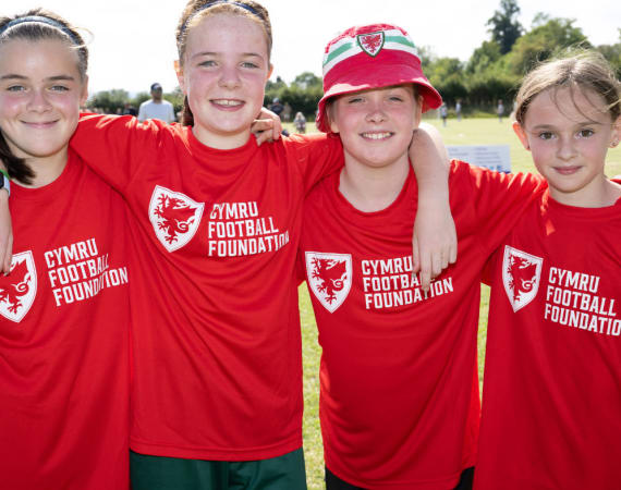 Four young girls wearing red Cymru Football Foundation shirts pose together outdoors on a sunny day, smiling at the camera.