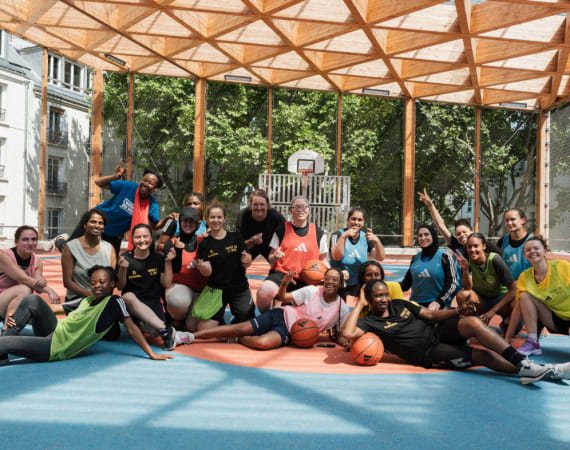 A diverse group of people pose and smile together on an outdoor basketball court under a wooden canopy, holding basketballs. Trees and buildings are visible in the background.