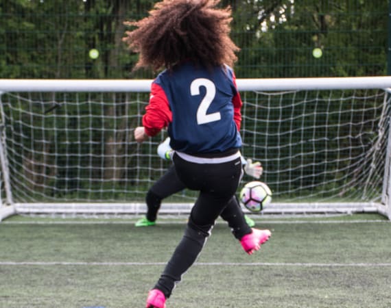 A soccer player wearing a blue jersey with the number 2 and pink shoes kicks a ball towards the goal, while a goalkeeper attempts to block the shot.