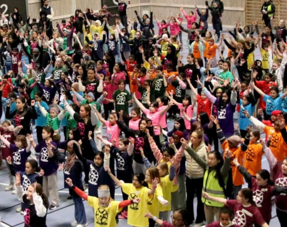 A large group of children wearing colorful shirts with a white logo stand in a gymnasium, raising their arms in unison. Some adults are present in the background.