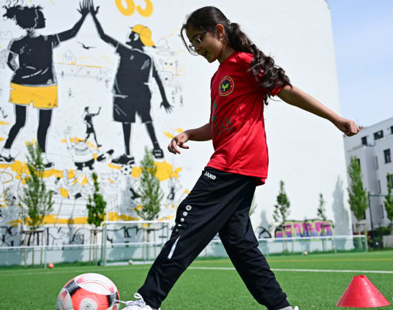 A girl in a red shirt and black pants practices soccer on a green field, preparing to kick a ball. Behind her is a mural of two children, one holding a soccer ball, on a white wall.