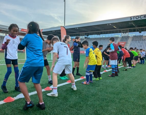 Children of diverse backgrounds stand in two lines on a soccer field, facing each other and participating in an activity. A stadium with empty seats is in the background.